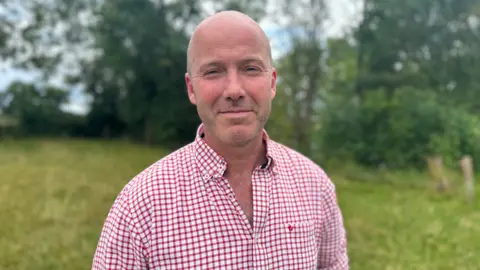 Charles Goadby is standing in a field. He is smiling and wearing a red and white checked shirt. 