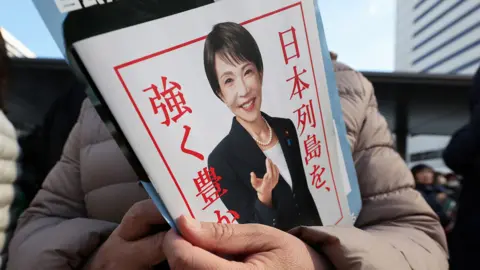 A close up of a pamphlet with a picture of Sanae Takaichi on the front held by a person off camera. Sanae Takaichi is wearing a black suit jacket, white t-shirt and pearl necklace in the photograph on the pamphlet. She is smiling. It has a white background and red border with red writing.