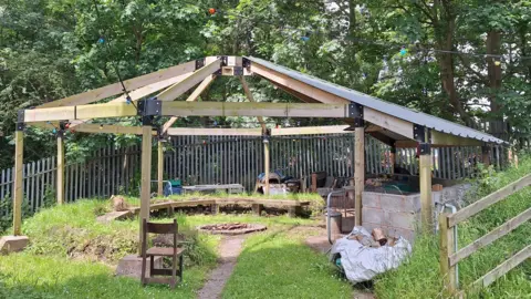 Gatis Community Space A wooden structure around a fire pit and there is a semi-circular wooden bench, a wooden chair and a path leading up to it as well as grass and fencing. 