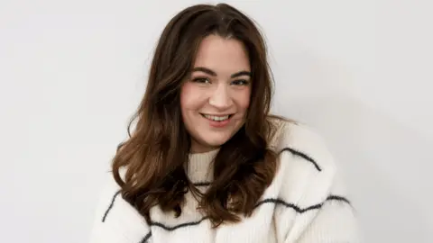 Abby Simms Abby Simms, from Nottingham City Council, pictured in a white jumper with thin black stripes. She has wavy brown hair and is smiling at the camera.