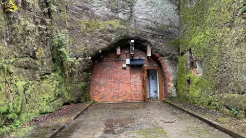 A mossy green rock face with a red brick building built into the side of it. There is a small brown door on the right hand side and path leading up to it.