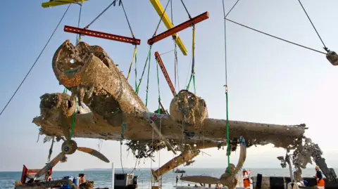 The shell of a large war plane that has been pulled from the sea bed. It is covered in brown algae, sand and debris. There are holes in its body and it is being hoisted up into the air by ropes