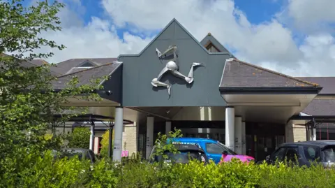 The front of Noble's Hospital has a large silver triskelion symbol above the entrance. Cars are parked in front and there is a green hedgerow in the foreground and a tree to the left.
