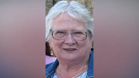 Meg Macdonald has short white hair and thin-framed glasses.