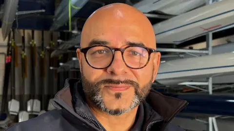 Omar Terywall, centre frame and looking directly out of the image. He is smiling slightly, has a dark, neatly trimmed beard, flecked with grey on his chin, and is wearing circular, black-framed glasses. He is bald. He is wearing a dark blue, collared top, but only the top of one shoulder on the left can be seen. Behind Omar are several sporting rowing boats mounted on racks inside a boathouse, along with rows of oars hung vertically along part of the wall behind him.