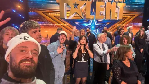 Whittingham Marketing & Consultancy A bearded man in a white beanie hat standing alongside a group of people on a stage.