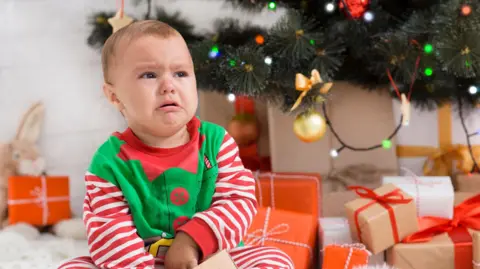 Getty Images A generic image of a grumpy-looking baby dressed in an elf onesie - in red and white stripes, green body and red buttons, sits in front of a Christmas tree and a pile of presents