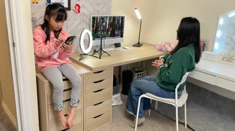 Moon talks to a camera as her daughter sits on a table beside her