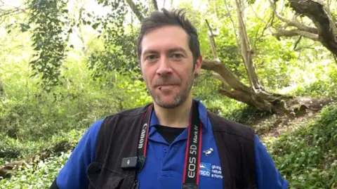 Rob Carmier is seen from his shoulders up standing in a sunny forest with green trees behind him. He is wearing a blue T-shirt, black vest and a camera around his neck.