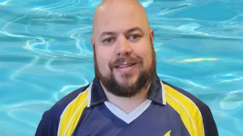 Rich Smith A man is by the water in his blue and yellow kit. He has a bear and is smiling at the camera.
