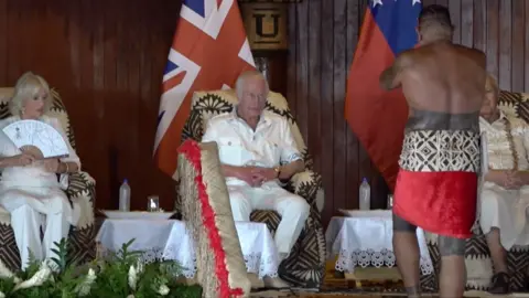 On a visit to Samoa, the King wears a white suit and is seated in front of a Samoan in traditional dress