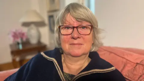 A grey haired woman wears red glasses as she sits on a peach coloured sofa. Over her right shoulder is a brown wooden table with a pink flower and white table lamp on it. There are two framed pictures on the wall beside the wooden table. The women is wearing a navy quarter zip sweater with a green t-shirt underneath. Around her neck is a small think gold necklace. 