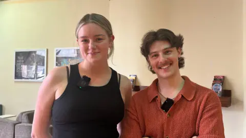 Isabella Verona/BBC Two people standing indoors in a foyer area with sofas and posters in the background. Both are wearing clip-on microphones. One person is wearing a sleeveless black top and the other is wearing a short-sleeve shirt.