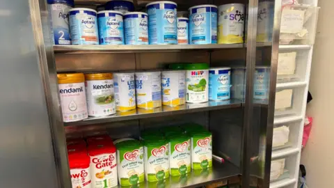 Megan Sinden Three metal shelves are lined with colourful tubs of baby milk. There is a tall, white set of drawers on the right with paper taped on each drawer denoting what is in it.