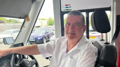 John Devine/BBC Joe Duffy has short brown hair and is sitting in the driving seat of a mini bus, he has a white short sleeve shirt on, and cars can be seen in the background.
