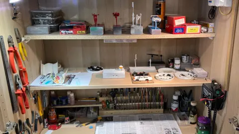 BBC/Jack Hadaway-Weller A compact workshop shelf is filled with model‑making tools, paints, brushes, and small parts neatly arranged.
Miniature car bodies, stands, and boxed kits sit alongside clamps, pliers, and precision instruments.
The setup suggests a dedicated, well‑used space for detailed scale model construction and finishing.