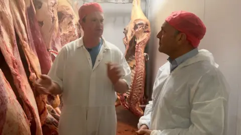 BBC/James Ingham Two men, wearing white overalls and hair nets, stand in the chiller section of an abattoir, carcasses hanging from rails all around them.
