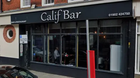 The bar has a black facade with a white sign. It has large square windows and its phone number on the top right of the building.