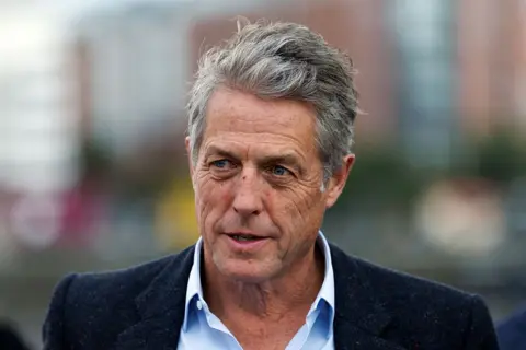 Getty Images Hugh Grant wearing a black jacket with a light blue open collared shirt.