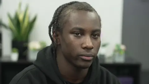 A close up of Chavez who is sitting down - he has braided hair and is wearing a black hoodie. There are some plants in the background. 