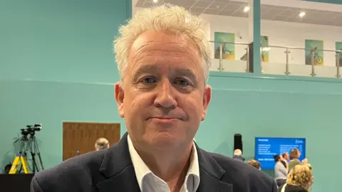 Charlotte Rose/BBC Paul Dundas is wearing a suit on election night. He has grey hair and is standing in a sports hall.