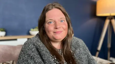 Louise is sat on a sofa in her living room. She has long, dark hair and is smiling at the camera. She is wearing a black and white patterned dress with a grey cardigan.