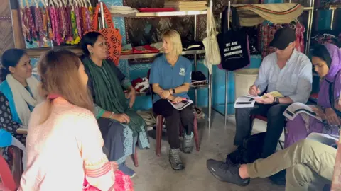 UNHCR A small group of people sit in a circle inside a room decorated with colorful fabrics and woven items. Amber wears a blue shirt with the UNHCR logo and holds papers, while another person is writing in a notebook. Shelves in the background display handmade crafts, and a black tote bag with white text reads, “WE ARE ALL ALIENS SOMEWHERE.”
