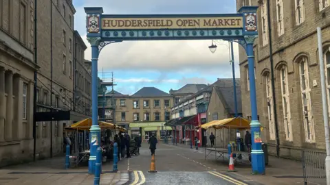 Olivia Courtney-Ashton/BBC York stone buildings tower above iron pillars and a gateway sign that reads Huddersfield Open Market. Beyond the archway, traders under yellow canopies line streets.