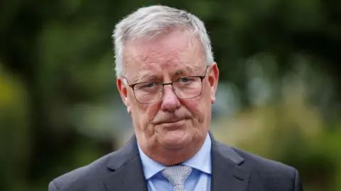 PA Media Nesbitt looking at the camera. He is wearing a grey suit jacket, pale blue shirt and a patterned cream and blue tie. He is wearing black framed glasses and has grey hair.