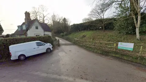 Google A lane with a cattle grid leading to a cream two-storey house. There is a white van parked at the entrance to the lane. A green sign on a fence reads: "Minstead Trust" and "Minstead Lodge"