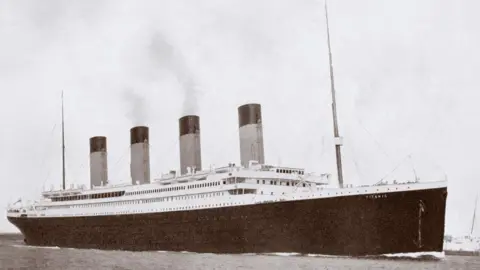 A black and white photo of RMS Titanic, a large ocean liner with four funnels, a black hull and a white upper deck. There are masts at the front and rear.