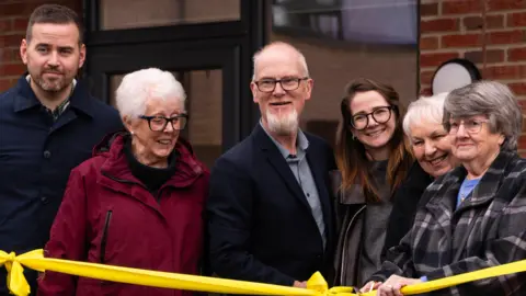 Basildon Council Six people standing behind a yellow ribbon. They are all smiling and standing in front of a building. One person has scissors poised ready to cut the ribbon.