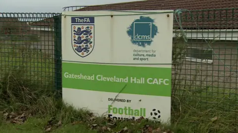 A large sign on a green metal fence that says 'Gateshead Cleveland Hall CAFC' in the middle, in white font with a green background. Above and below this, the background of the sign is white. In the top left is the FA logo and in the top right is a logo that. says 'dcms', 'department for culture, media and sport'. At the bottom right it says 'Delivered by Football Foundation' with their logo. There is grass either side of the fence, and a building behind the sign.