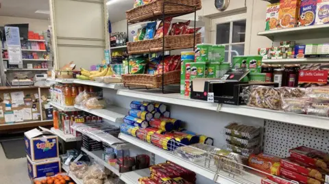 Rows of shelves containing biscuits, snacks and fruit, with a Post Office window at the end.