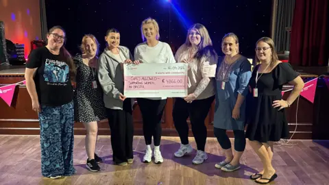 A group of seven women standing in a line inside a dimly lit hall. The three women in the middle are holding a large cheque for £4,866.