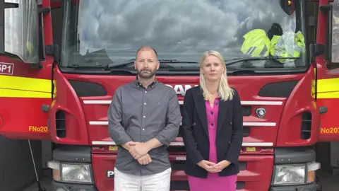 LDRS / Rebecca Paul A man and a woman stand in front of a fire engine at Banstead Fire Station. They have neutral facial expressions.