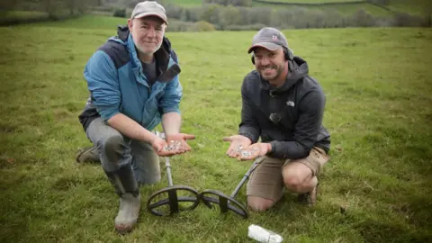 Michael Eakers/Brian Dixon Two people kneel in a grassy countryside field holding handfuls of freshly found silver coins, with metal detectors placed on the ground between them.