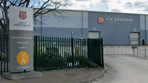 Google AV Dawson building at Middlesbrough port. It is a large grey industrial unit with a number of cream roller doors. To the left of the entrance road is a freestanding rectangular sign saying the Port of Middlesbrough and signposting the automotive metals story, the HGV entrance and offices. In front is green metal fencing, two open gates and the kind of raisable barrier you have at level crossings - a white bar with sections hanging beneath.