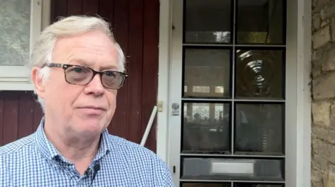 Andy Baker has short white hair and slightly darkly tinted glasses. He is wearing a blue and white checkered button shirt. He stands by the front door of his house in the left hand side of the frame, looking out of frame to the right. 