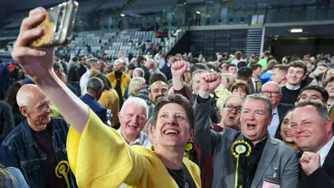 Alison Thewliss - a woman wearing a yellow jacket and with short dark hair, takes a selfie of herself and dozens of celebrating SNP volunteers, on the floor of an election count 