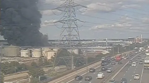 National Highways Traffic cam image of black clouds of smoke rising into the sky, near an oil terminal. Vehicles are passing along the motorway nearby