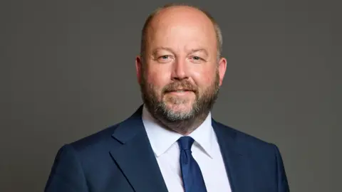 UK Parliament A portrait image on a grey background of Nick Timothy wearing a blue suit, white shirt and tie. 
