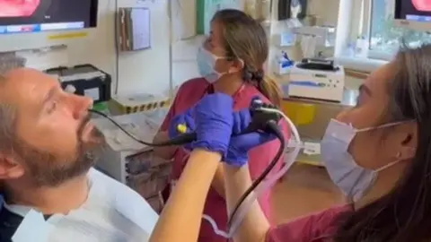 Shiying Hey David the patient sits in a chair with a tube inserted into his nose by the surgeon in raspberry scrubs as a nurse also in raspberry scrubs looks on