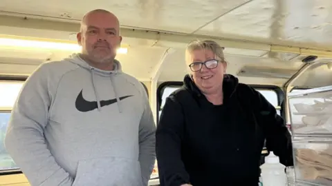 Lara King / BBC Mark De La Touch has a bald head and is wearing a grey hoodie. He is smiling and has his hands in his pockets. Clare De La Touche is standing next to him wearing a black top and black glasses. She is also smiling. They are standing next to plastic containers with plastic cutlery in them.