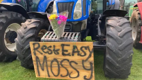 A sign that reads "Rest Easy, Mossy" is on the front of a tractor with a bunch of flowers in tribute to Harry.
