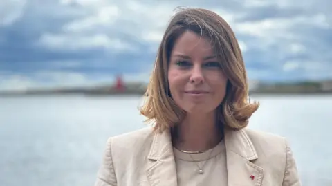 Kim McGuinness is standing with her back to the sea on a beach. She has blonde, shoulder length hair, is half smiling, and is wearing a cream top and cream jacket. In the background, out of focus, is a lighthouse on a pier jutting out into the water.