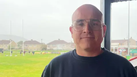 BBC Wales News Steelworker Adam Williams, with glasses and wearing a sweatshirt, and standing near a rugby field looks into the camera