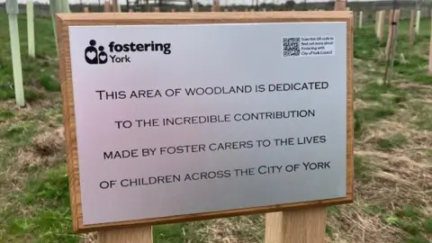 BBC/Katie Radley A plaque is mounted on a wooden frame, in the background is a field planted with trees. The plaque reads "this area of woodland is dedicated to the incredible contribution made by foster carers to the lives of children across the City of York. 
