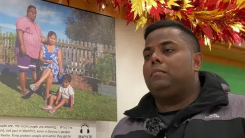 Mahi , stood in front of a picture of him, his wife and son.