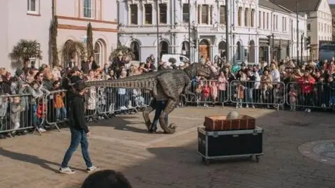 A dinosaur puppet with man with dark hat, dark top, blue jeans and trainers. They are surrounded by a metal fence with an onlooking crowd. There is a box of props in the foreground. They are on paving with buildings in the background.